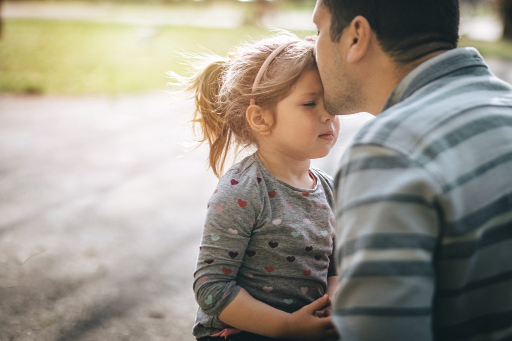 Young father sitting with his daughter in park and kissing her
